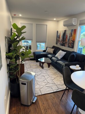 A modern living room with a black sofa, a round coffee table, a small side table, a potted plant, and a cityscape painting on the wall. - #2 Richards Ave Cozy Brand New 1BR apt half mile CASV (Oneonta)