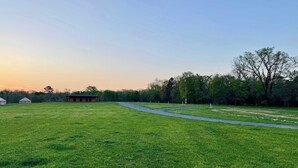 A grassy field with a gravel path, a small wooden building, and trees in the background. - RV site 1 @ The River Bluff Cabins (Tahlequah)