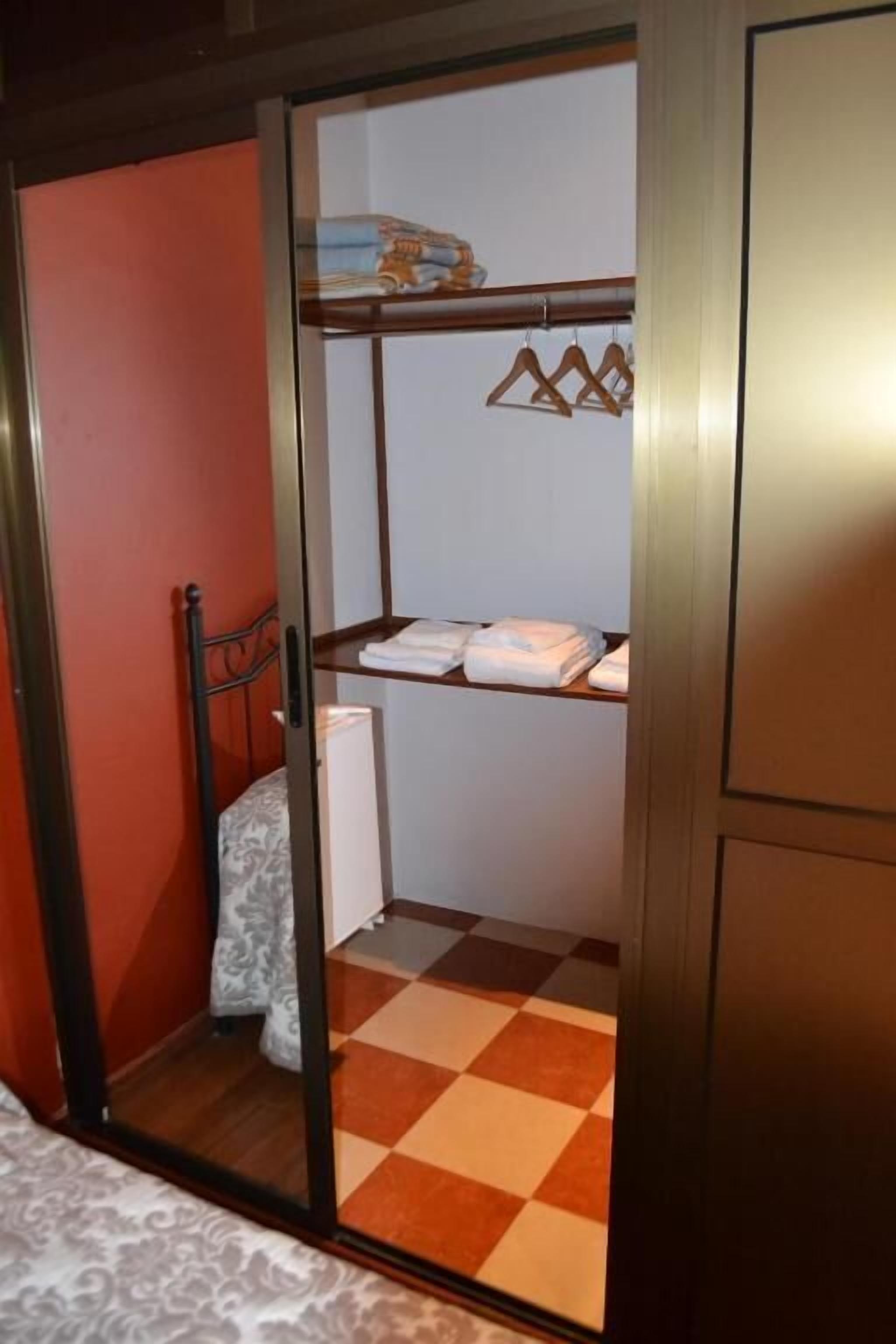 A room with a checkered floor, a bed, a wardrobe, and a closet with hanging clothes.