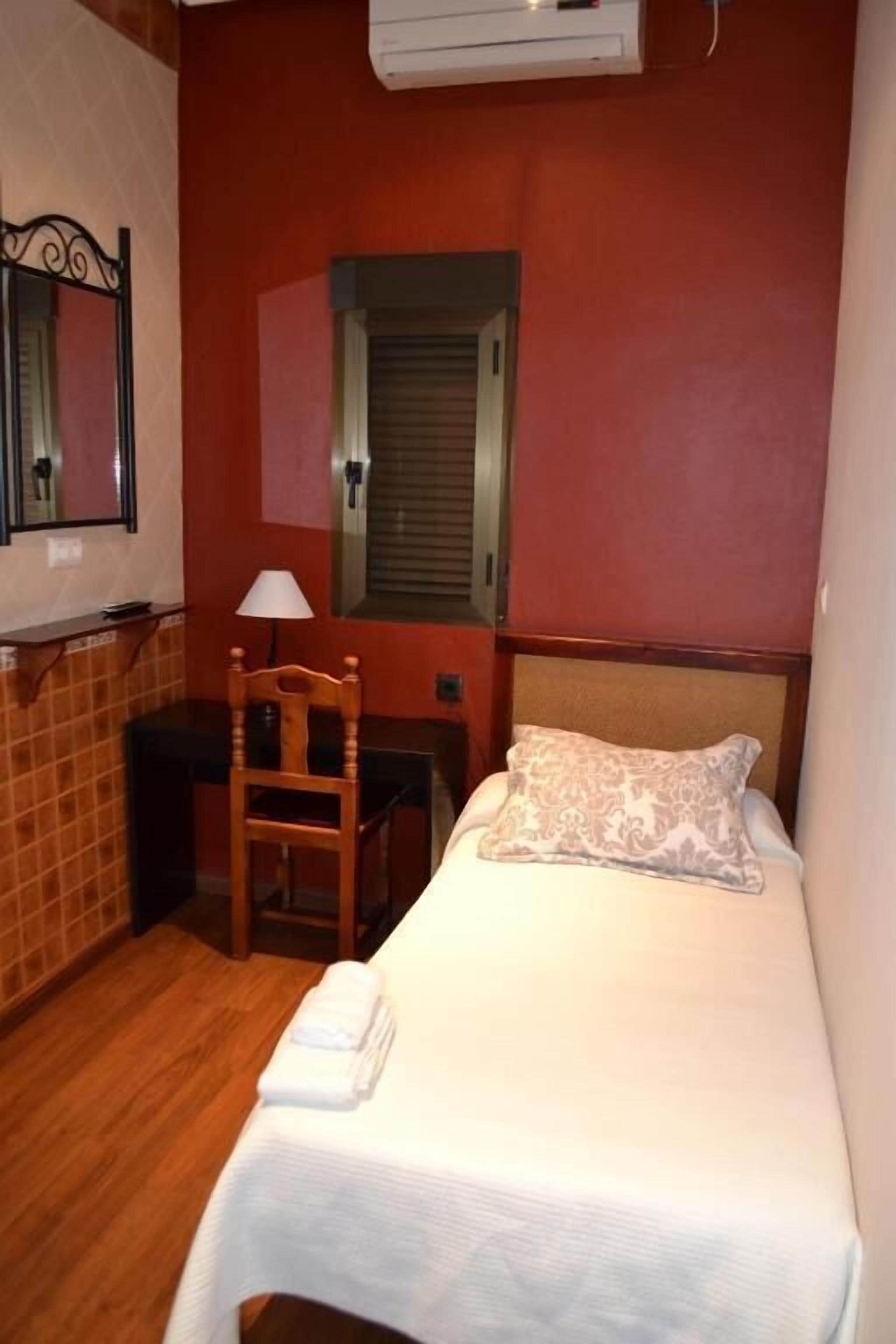 Una habitación de hotel pequeña con una cama, un escritorio, una silla, una lámpara y un espejo.
