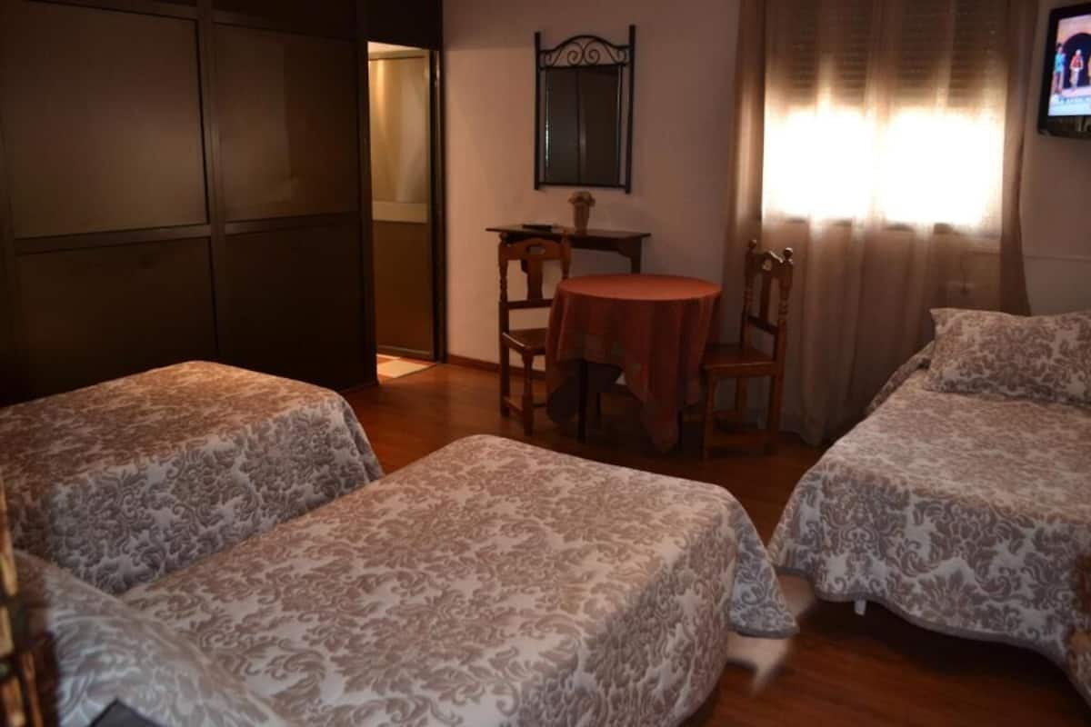 Ένα δωμάτιο με δύο κρεβάτια, ένα μικρό στρογγυλό τραπέζι με καρέκλες, έναν καθρέφτη στον τοίχο και ένα παράθυρο με κουρτίνες.