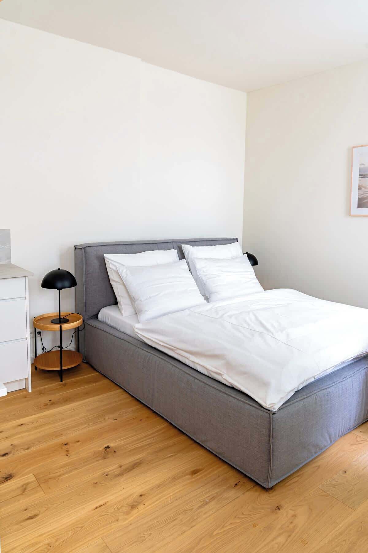 A modern bedroom with a grey bed, white bedding, a round wooden nightstand with a black lamp, and a white dresser.
