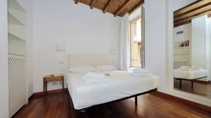 Una camera da letto con pavimento in legno, un letto con lenzuola bianche, un comodino, una finestra con tende e uno specchio appeso al muro.