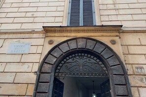 Une entrée en arc surmontée d’une grille en métal décorative, une enseigne indiquant « Vicolo delle Grotte », et une fenêtre avec des volets.