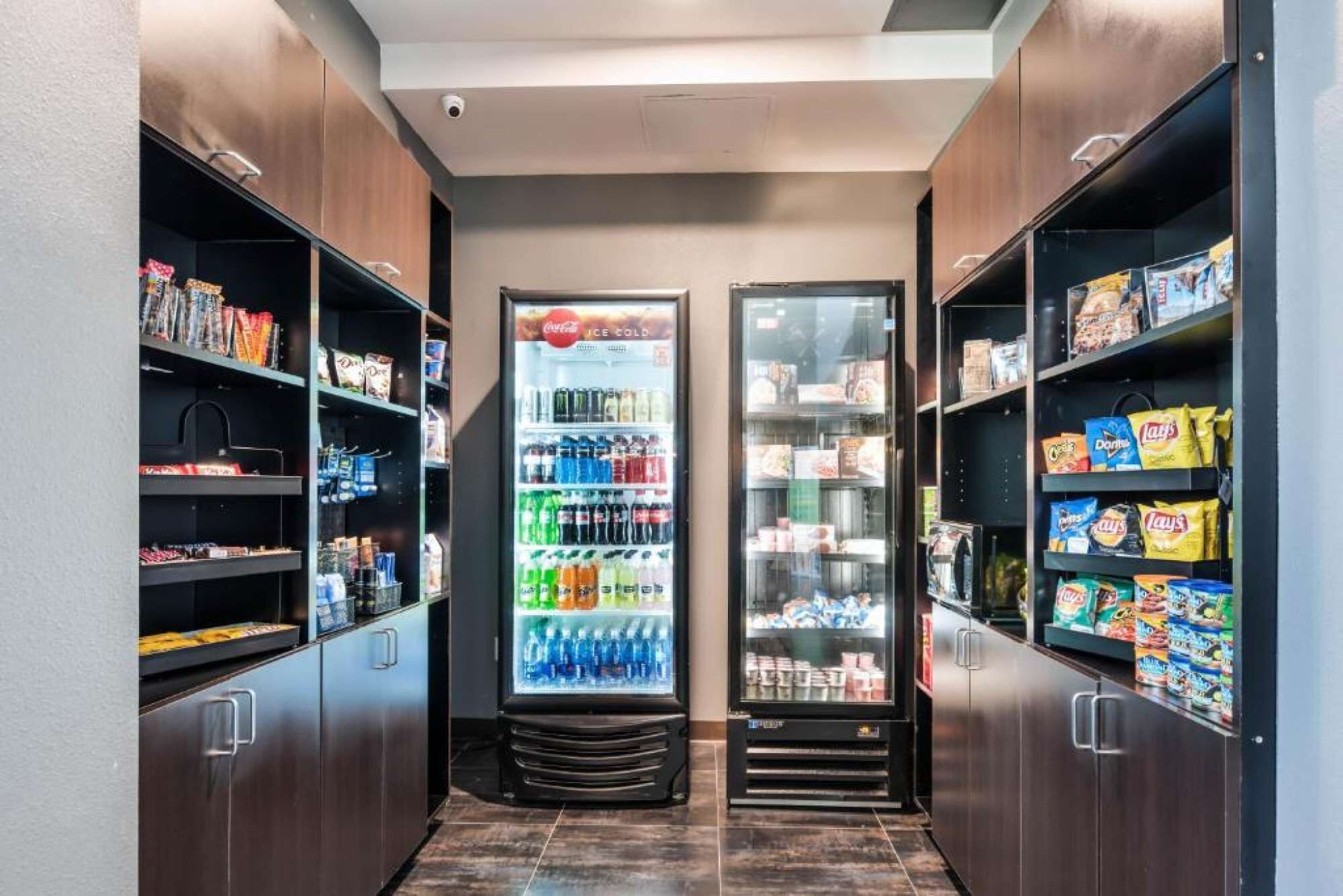 Un pasillo estrecho con dos máquinas expendedoras llenas de bebidas y aperitivos, flanqueado por estantes con diferentes productos envasados.