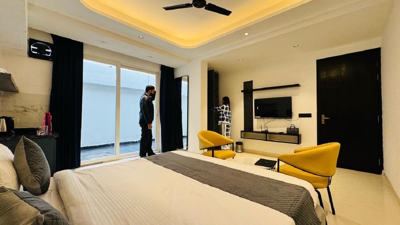Une chambre d’hôtel moderne avec un grand lit, deux fauteuils jaunes, un bureau, une télévision et une porte coulissante donnant sur un balcon.