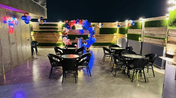 Un área de patio al aire libre con mesas redondas y sillas de mimbre negro, decorada con globos y un letrero de neón.