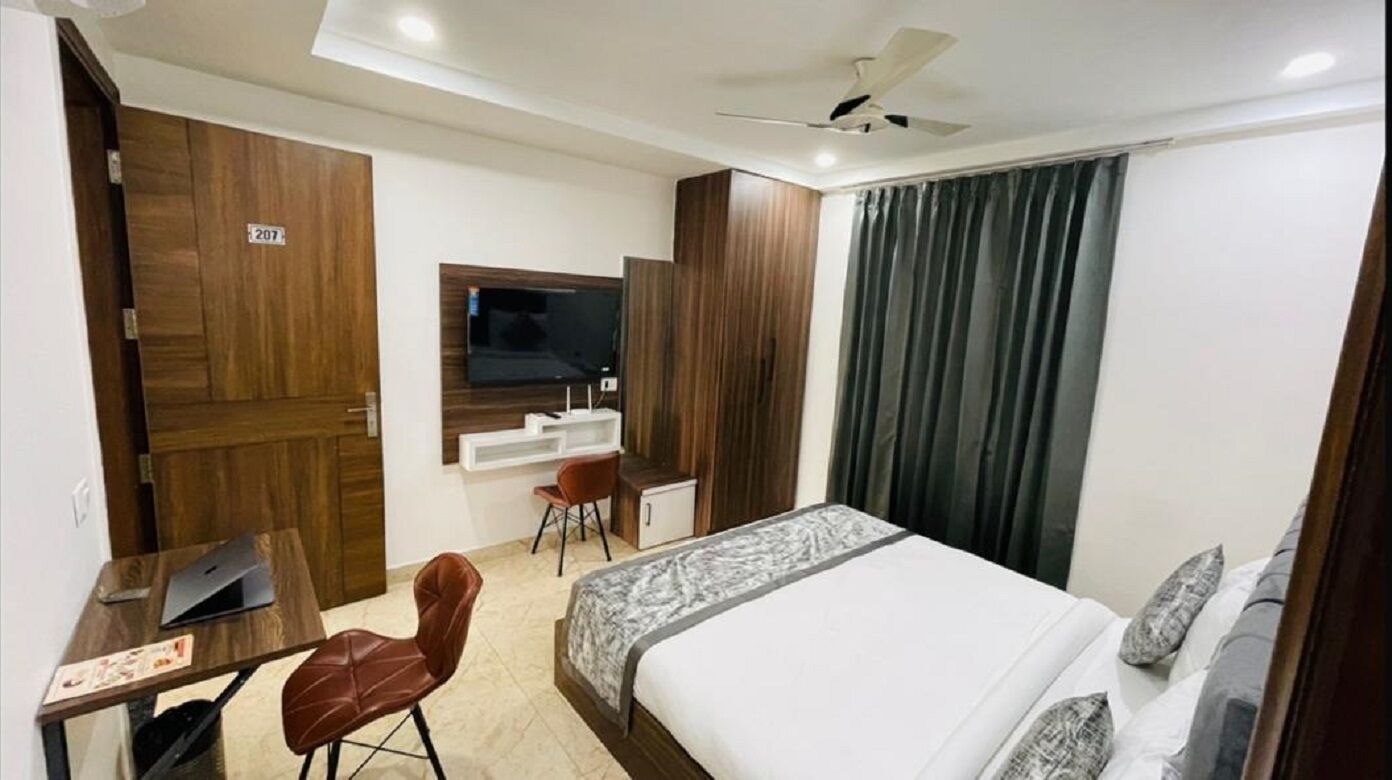 Ein Hotelzimmer mit einem Bett, einem Schreibtisch mit einem Laptop, einem Sessel, einem an der Wand befestigten Fernseher und einem Deckenventilator.