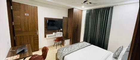 Ein Hotelzimmer mit einem Bett, einem Schreibtisch mit einem Laptop, einem Sessel, einem an der Wand befestigten Fernseher und einem Deckenventilator.