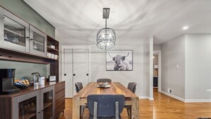 Een eetgedeelte met een houten tafel, stoelen, een koffiehoek en een aan de muur bevestigd schilderij.