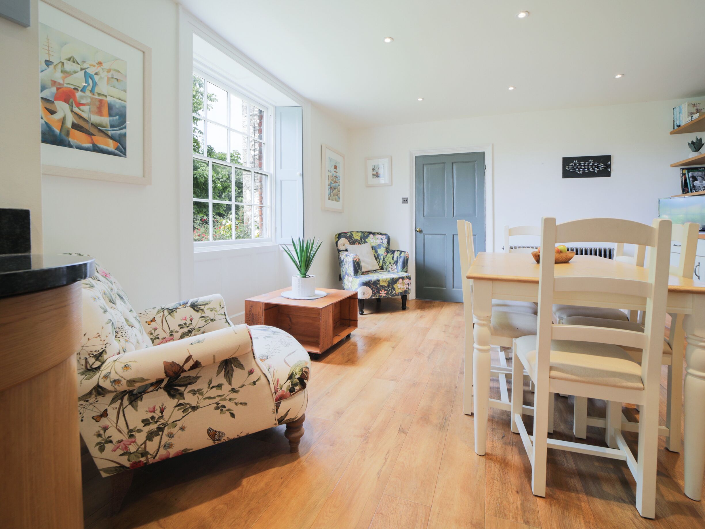 A spacious living room with a floral sofa, a wooden dining table with chairs, and a large window providing natural light.