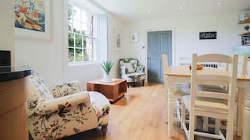A spacious living room with a floral sofa, a wooden dining table with chairs, and a large window providing natural light.