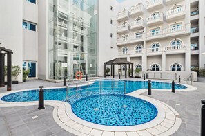 A modern hotel with a central swimming pool, a glass facade, and a multi-story building with balconies. - Modern 1BR in JVC, 2 Baths, Pool & Gym, Fully Equipped Kitchen (Dubai)