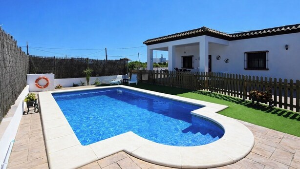 Una piscina con borde curvo, un edificio blanco con tejado de azulejos y una valla de madera.