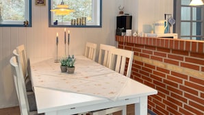 Un coin repas avec une table blanche dressée pour quatre personnes, entourée de chaises blanches. On peut voir trois bougies sur la table et une petite plante en pot au centre. Un mur de briques et une fenêtre donnant sur des arbres sont visibles à l’arrière-plan.