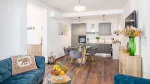 A modern apartment with a living room, dining area, and kitchen. There is a blue sofa, a dining table with chairs, and a kitchen with a stove and sink.
