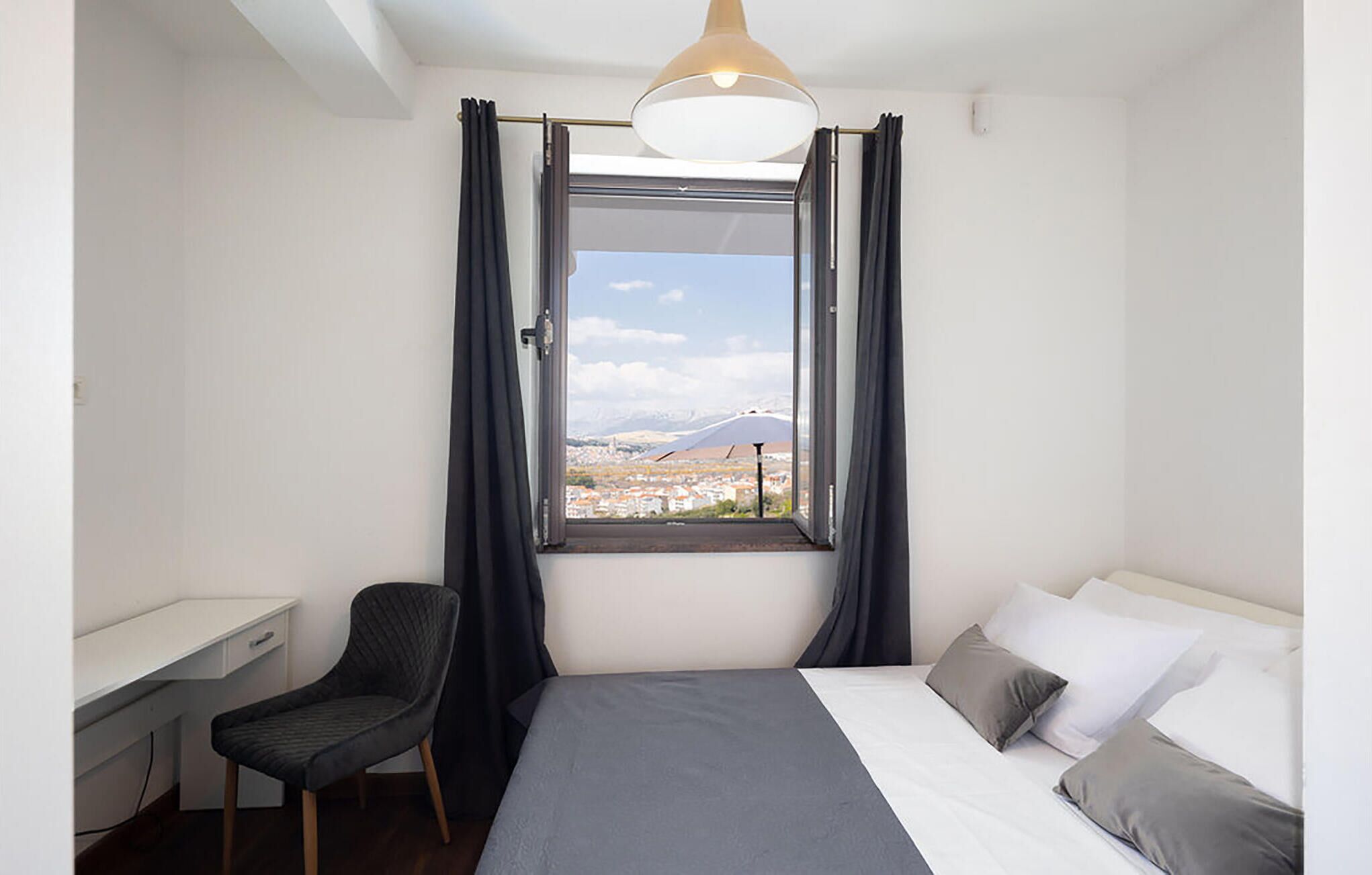 A bedroom with a bed, a desk, and a chair. There is a window with curtains, and a view of a cityscape and mountains.