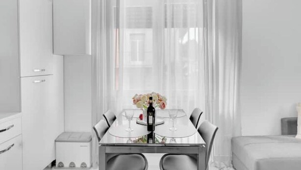 Une chambre d’hôtel moderne dotée d’un coin repas comprenant une table en verre, des chaises blanches et une bouteille de vin.