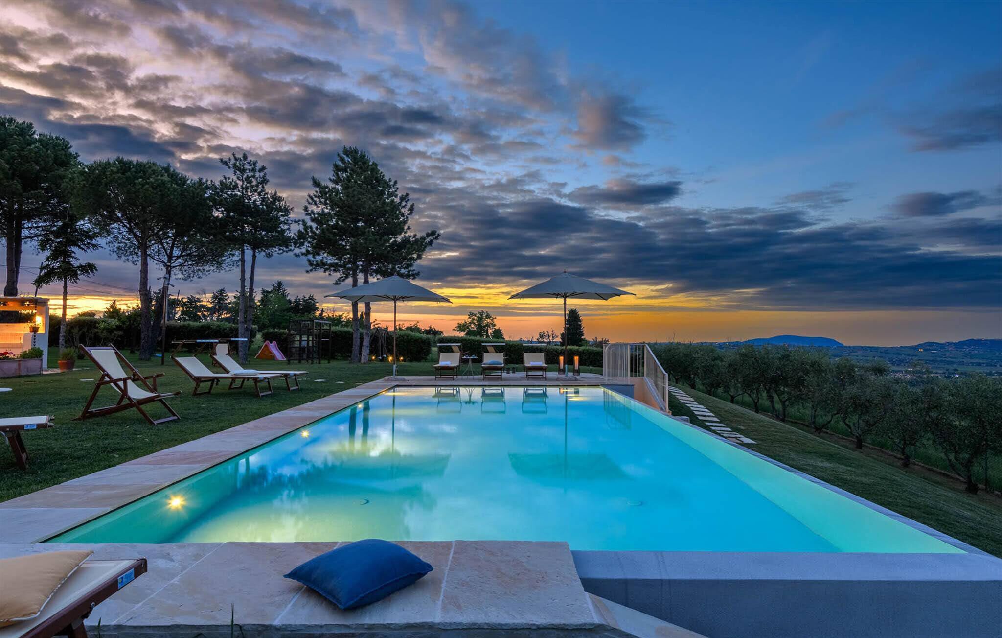 Una piscina con lettini e ombrelloni, circondata da alberi e vista sul tramonto.