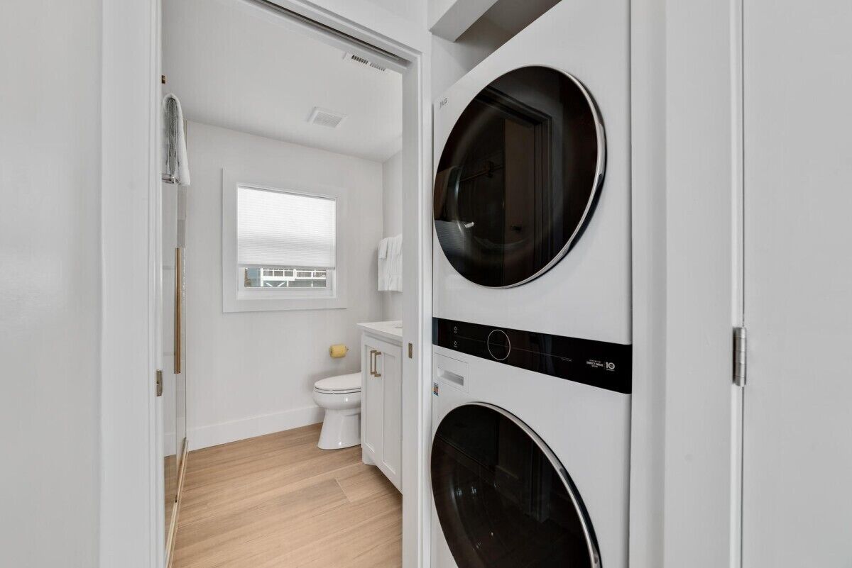 A compact laundry room with two front-loading washing machines, a white toilet, and a window above the sink.