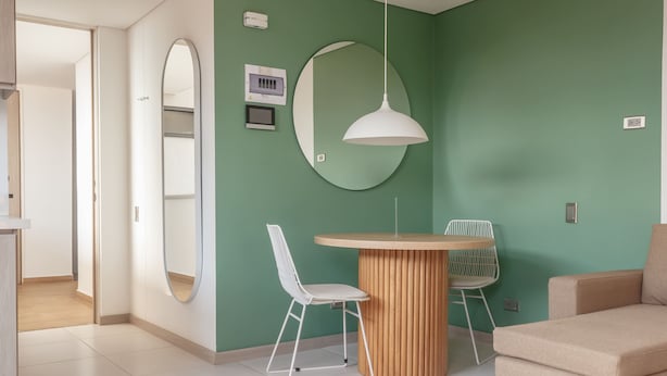 A modern living room with a round wooden table, two white chairs, a beige sofa, a large mirror, and a pendant light. - NOK Stylish Studio w Balcony in Laureles Medellin (Medellín)