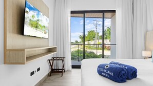 A hotel room with a large bed, a mounted TV, a wooden shelf, and a view of the outdoors. - NOK Modern Apartment The Towers at Vistacana Punta Cana (Punta Cana)