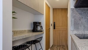 Una cocina estrecha con una cafetera en un estante, una puerta de madera y un cubo de basura.