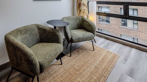 Un espacio de oficina moderno con dos sillones verdes, una mesita redonda, un cuadro enmarcado en la pared y vistas a un edificio de ladrillo a través de amplias ventanas.
