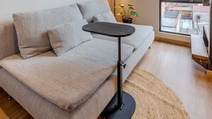 En moderne stue med en grå sofa, et lille rundt bord, en potteplante og et stort vindue med udsigt over byen.