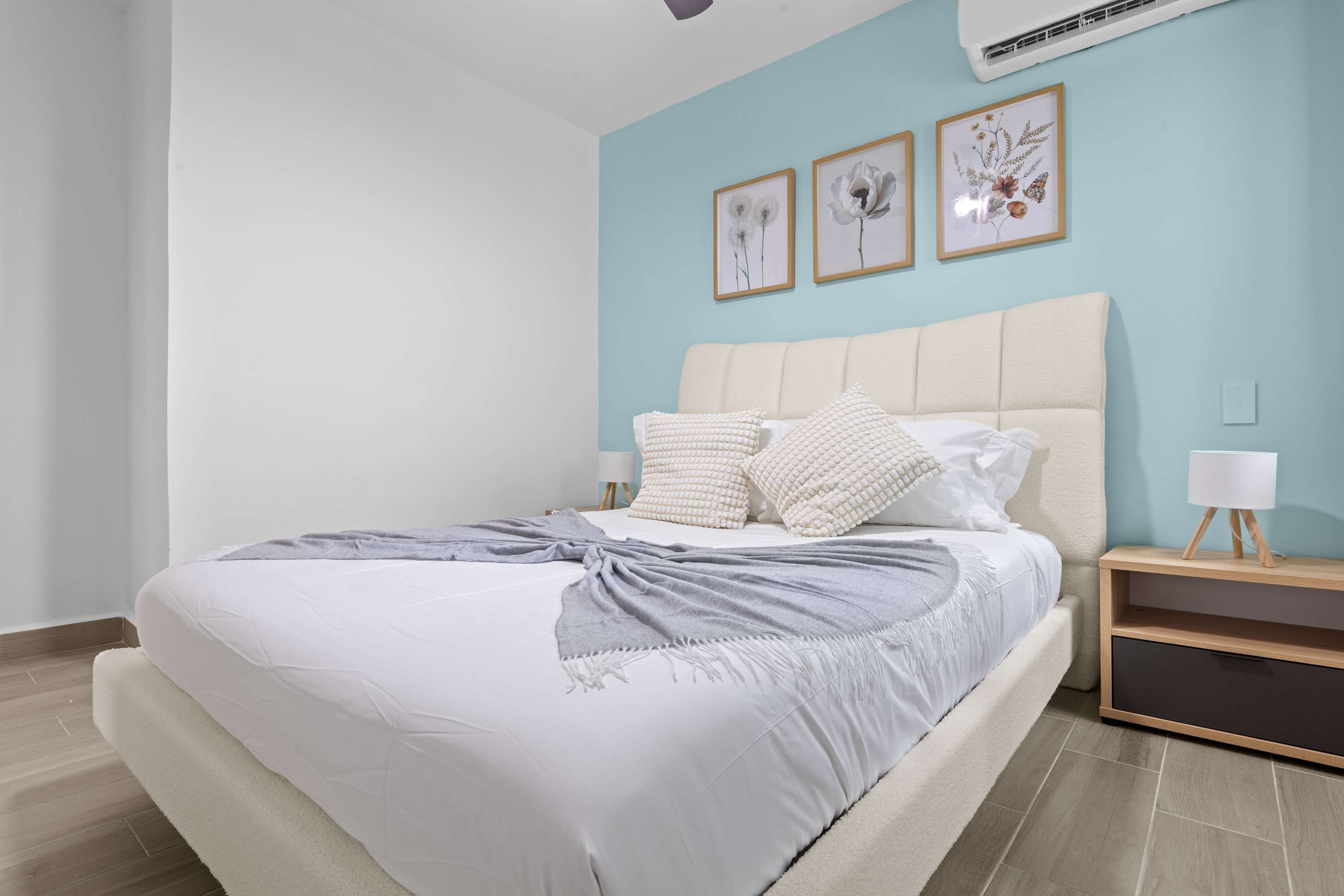 A neatly made bed with a grey and white duvet, a cream-colored headboard, and a bedside table with a lamp.