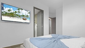 A bedroom with a bed, a TV on the wall, and a door leading to another room. - NOK Designer 2BR Loft Jardines 3 Punta Cana (Punta Cana)