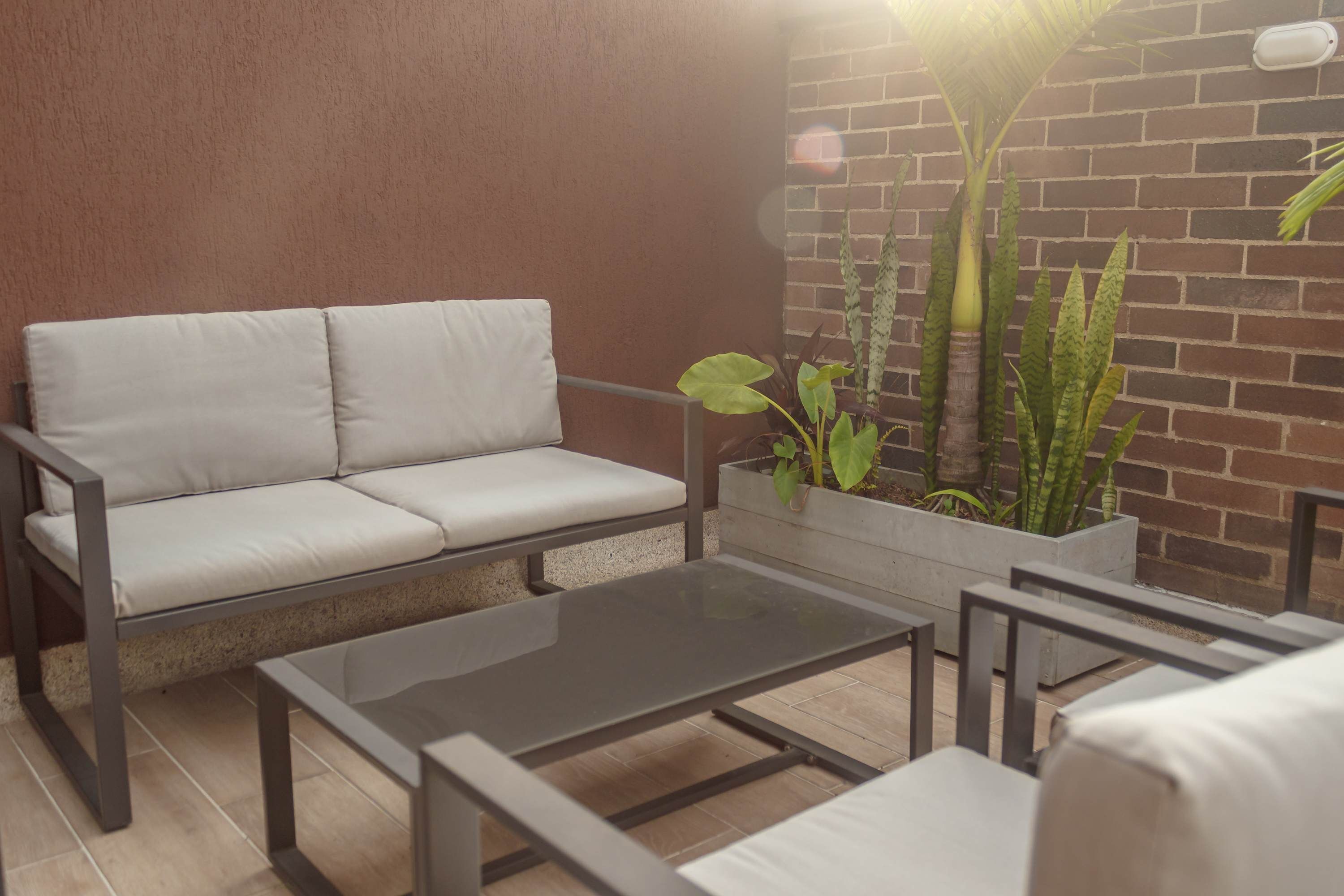 A modern outdoor seating area with a sofa and a glass table, surrounded by potted plants and a brick wall.