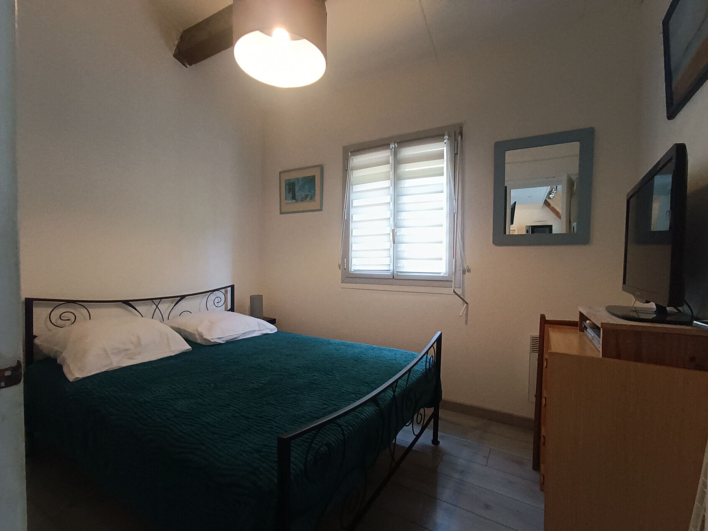 Une chambre à coucher avec un lit, une fenêtre avec des stores, un miroir et un téléviseur posé sur un meuble en bois.