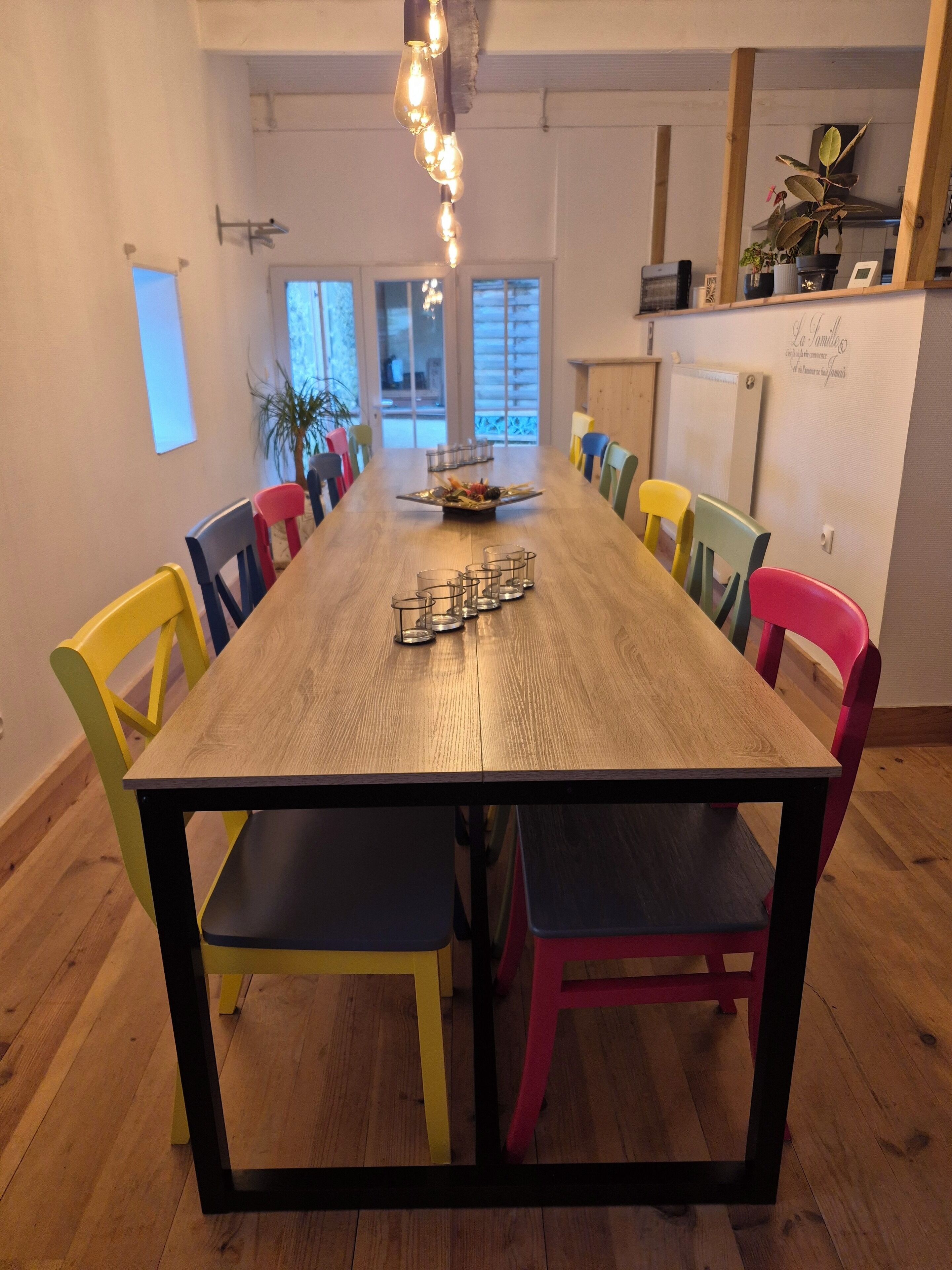 Un comedor con una larga mesa de madera, sillas coloridas y una lámpara decorativa en el centro.