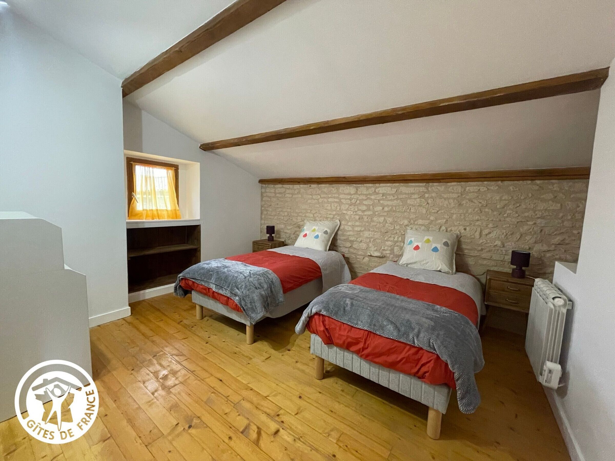 Een slaapkamer met twee bedden, een stenen muur, houten balken, een raam met gordijnen en een radiator.