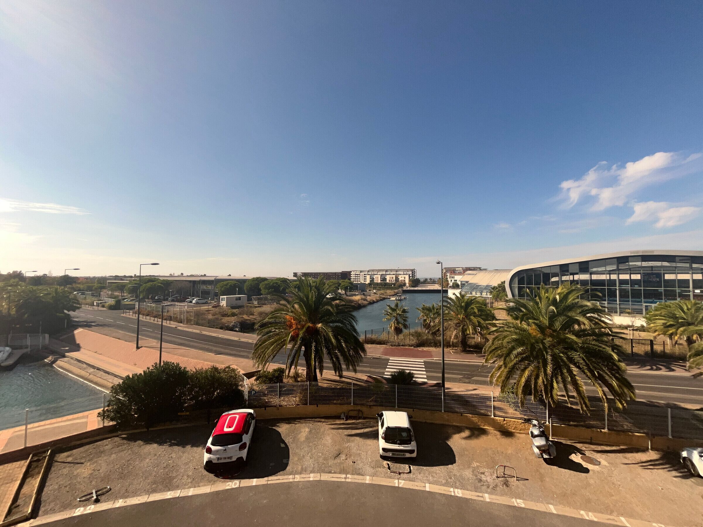 A parking area with a white car and a white van, palm trees, and a modern building with a curved roof.
