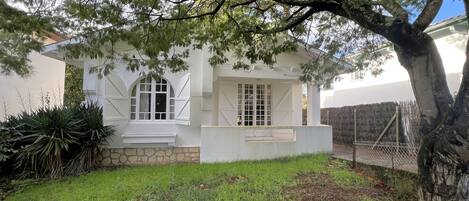 A white building with a stone base, a small porch, and a fenced yard with grass and plants.