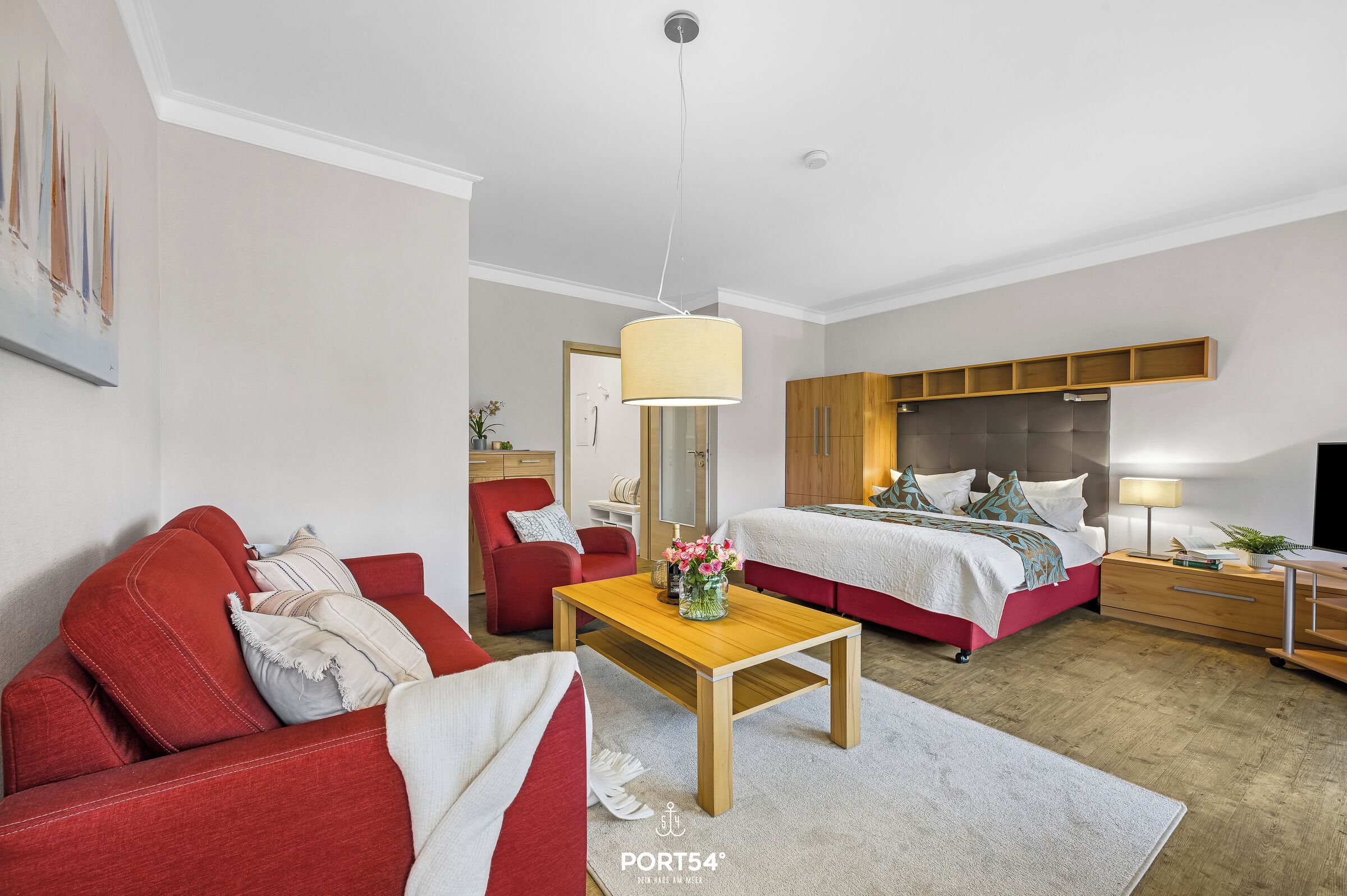 Habitació d'hotel moderna amb un sofà vermell, una taula de centre de fusta amb flors, un llit amb capçal i un escriptori amb una làmpada.