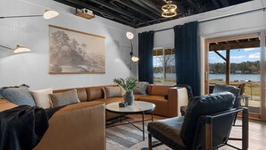 Ein modernes Wohnzimmer mit einem Ecksofa, einem runden Couchtisch und einem Ledersessel. Ein großes Fenster bietet Blick auf einen See.