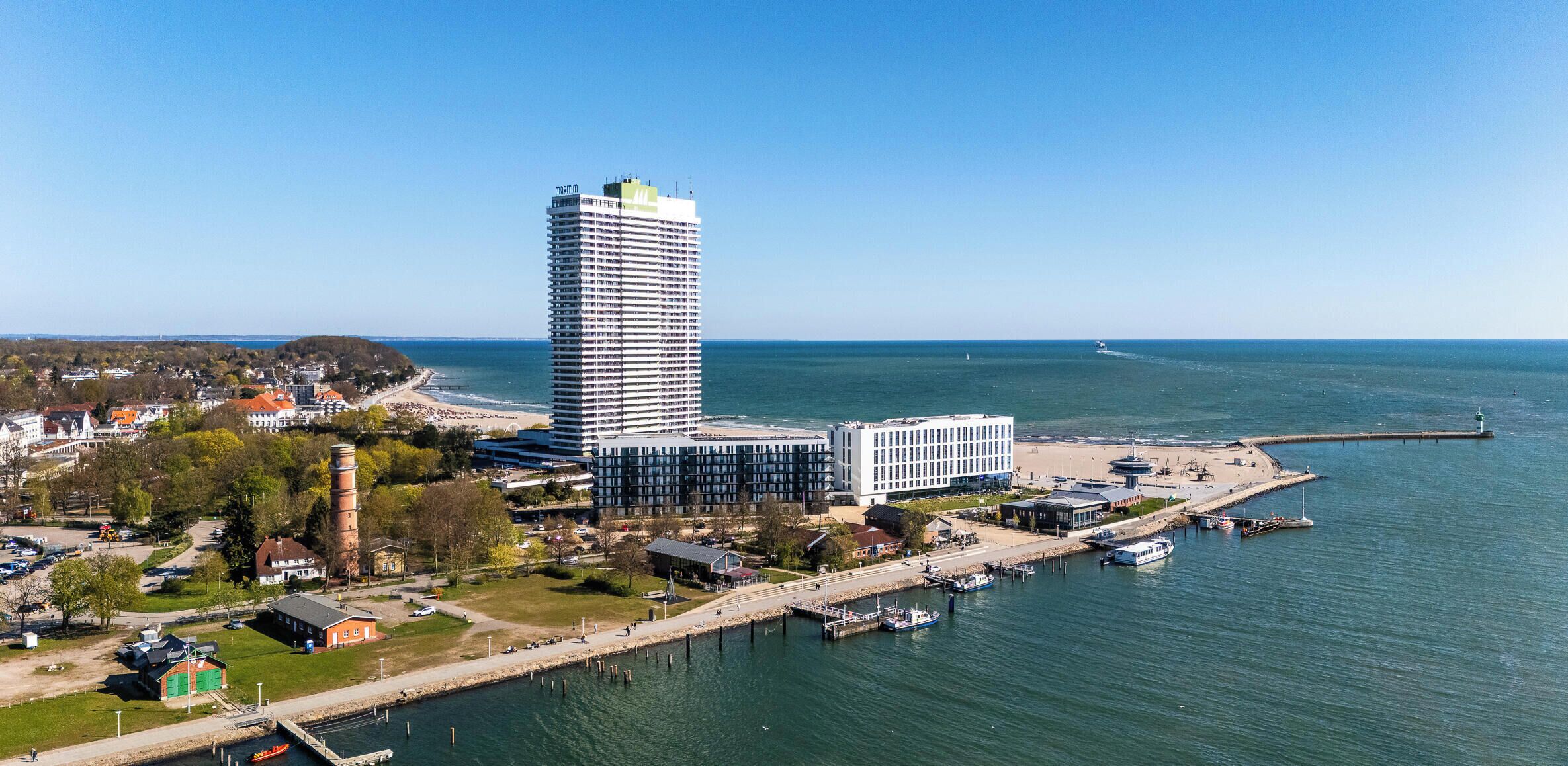 Eine Küstenstadtlandschaft mit einem markanten Hochhaus, einem Yachthafen mit Booten und einer Strandpromenade.