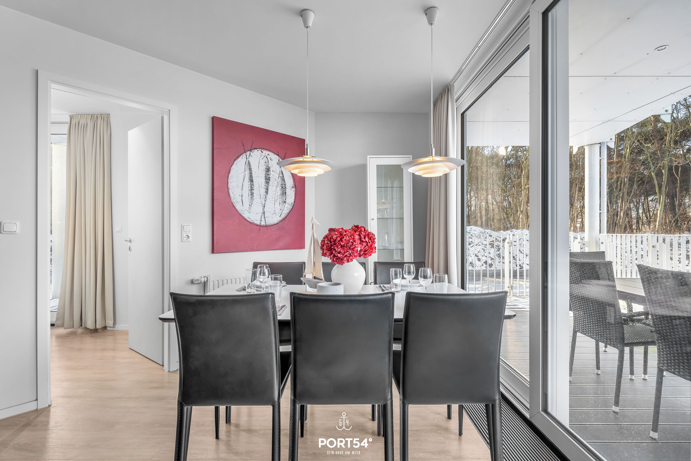 Ein moderner Essbereich mit einem großen Tisch, gedeckt für vier Personen, schwarzen Stühlen und einer Vase mit roten Blumen. Ein großes Fenster bietet einen Blick ins Freie.