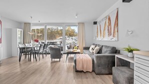 A modern living room with a grey sofa, a dining area with a table and chairs, and a large window with a view of trees and hills. - Bright Family Apartment with Covered Balcony and Wellness near Priwall Beach (Lübeck)