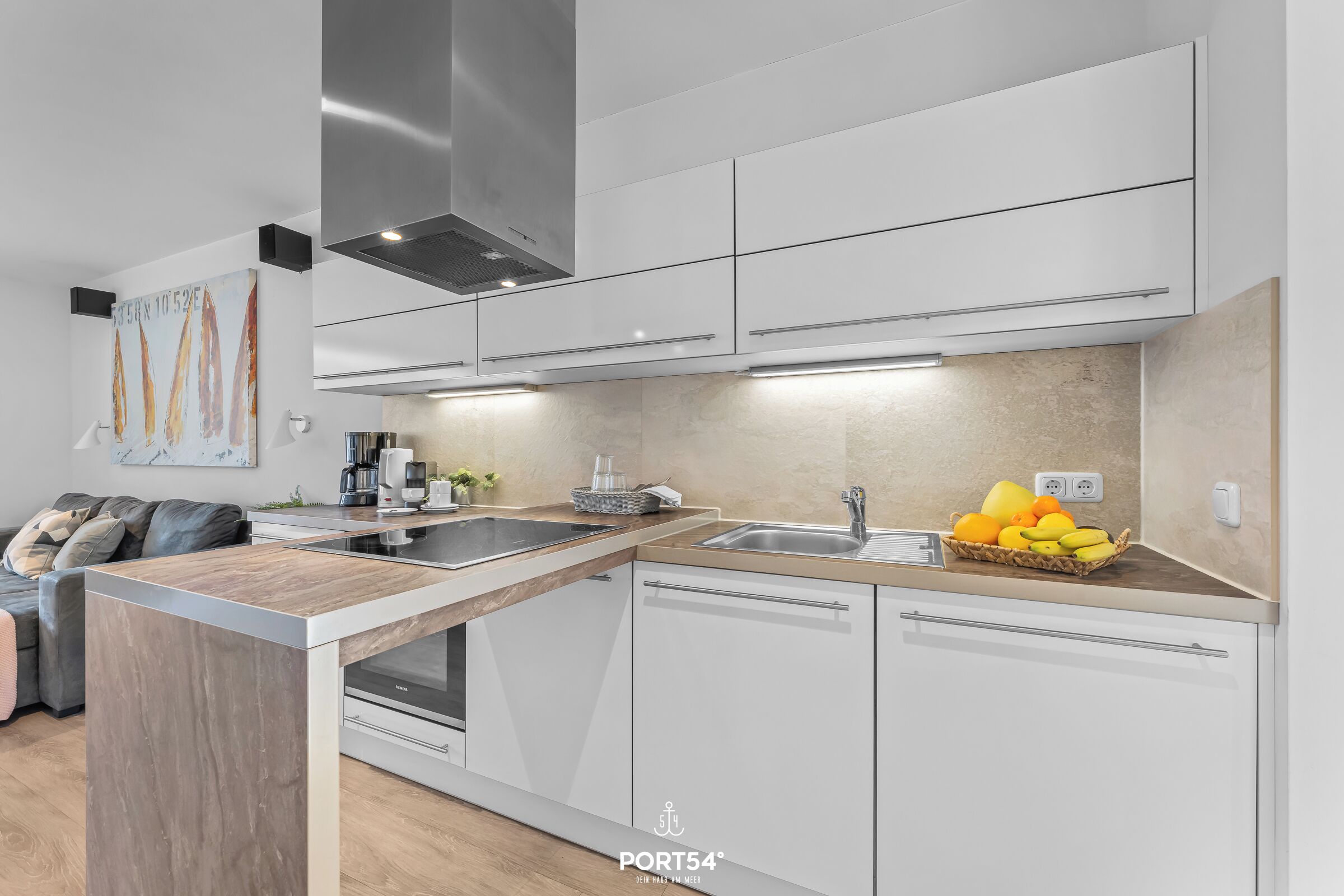 Eine moderne Küche mit weißen Schränken, einer Holz Arbeitsplatte und einem eingebauten Backofen. Auf der Arbeitsplatte steht eine Obstschale und ein Kaffeeautomaten.