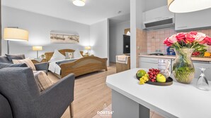 Ein modernes Hotelzimmer mit einem Bett, einer Sitzecke mit Sofa und Sesseln, einer Küchenzeile mit Esstisch und einer Vase mit Blumen auf dem Tisch.