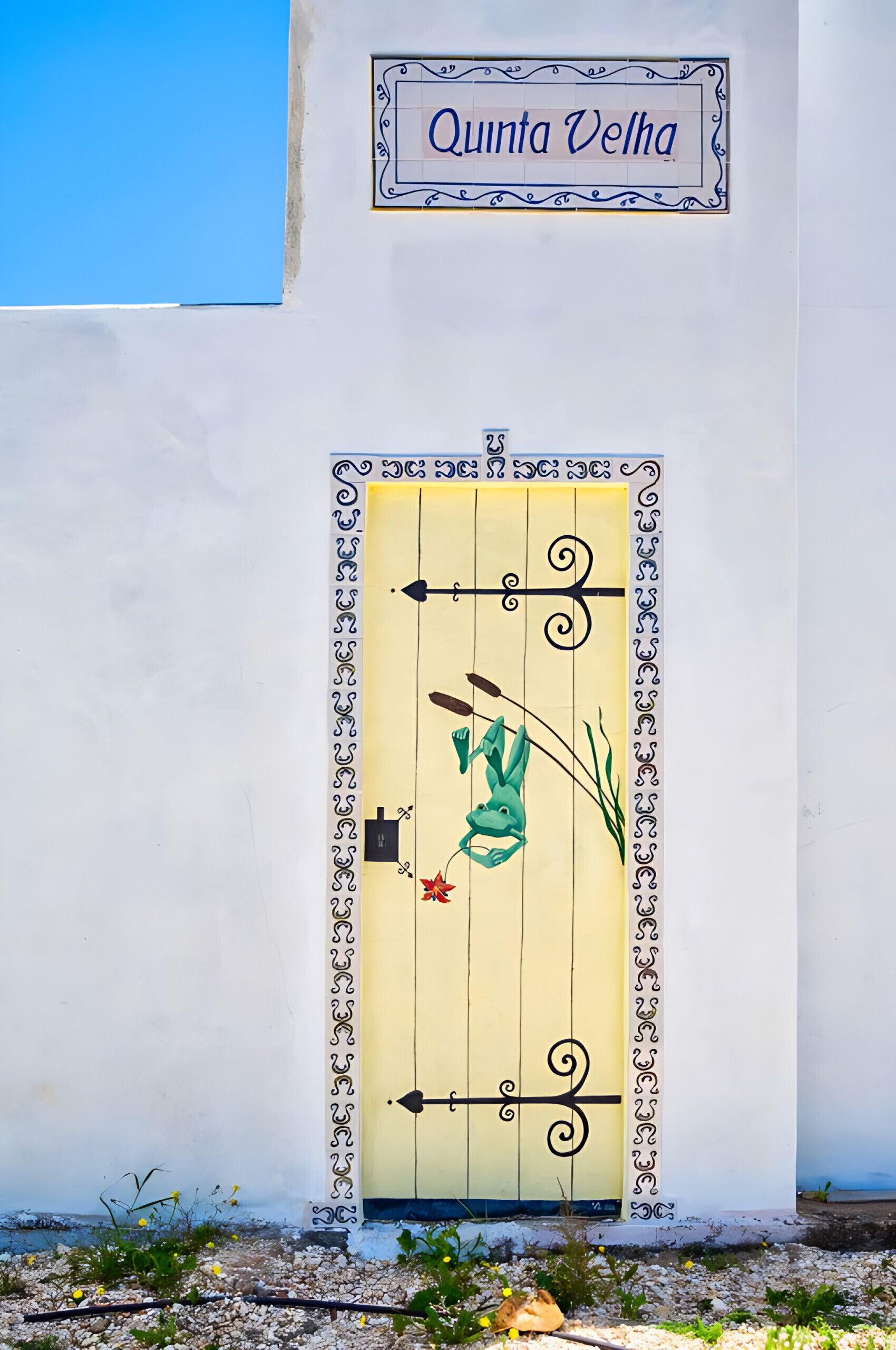 A white wall with a sign reading "Quinta Velha" above a decorative door with a yellow panel and black ironwork.