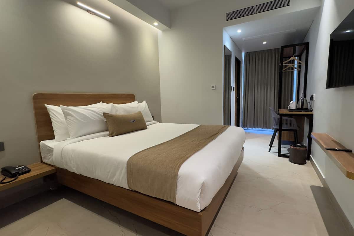 A modern hotel room with a wooden bed, white linens, a bedside table with a phone, a desk with a chair, and a flat-screen TV mounted on the wall.