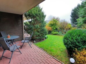 Un patio con mesa y sillas, un sendero de ladrillo y un jardín bien cuidado con diversos arbustos y árboles.