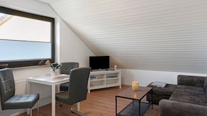 Un dormitorio tipo loft moderno con techo inclinado, un ventanal grande, un televisor sobre un mueble blanco, un sofá y una zona de comedor con sillas.