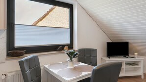 Una habitación con una mesa de comedor, sillas, un televisor sobre un mueble, una ventana con persianas y una planta.