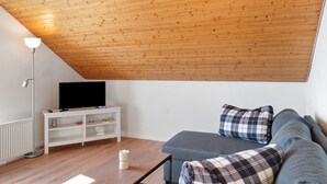 En stue med træloft, et fjernsyn på et hvidt stativ, en blå sofa og en gulvlampe.